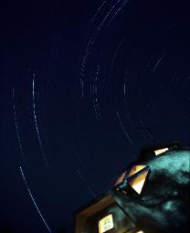 A four-hour time exposure photograph of the night sky showing star trails and with a geodesic home in the foreground.